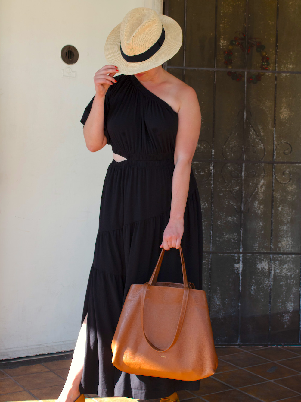 A woman with a hat tilted across her face, wearing a one-shoulder Cuyana dress, and holding a leather tote bag.