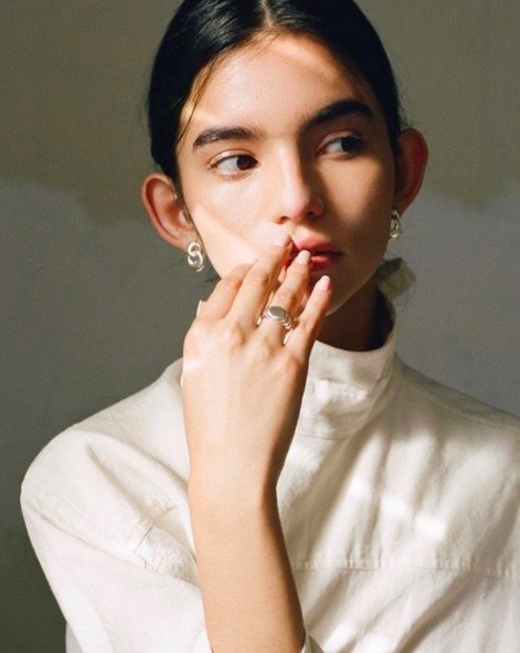 A model wearing a white turtleneck with black hair pulled back and their fingers on their lips, wearing a chunky silver ring and double loop silver earrings.