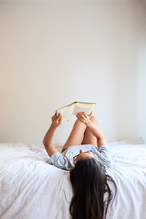 Beautiful pretty woman in glasses lying with novel on bed at home.